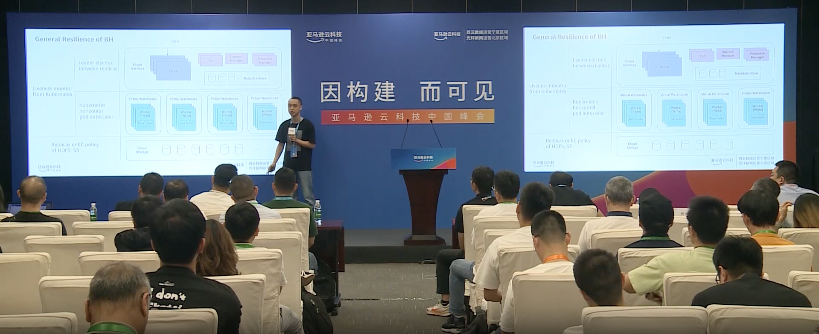 A Chinese man speaking with a presentation playing behind